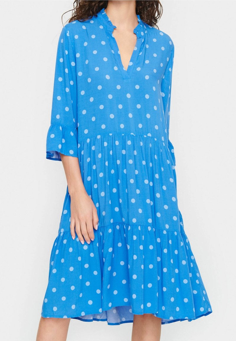 Blå prickig klänning i lättviktig tyg, har V-ringning, volangärmar och en flerskiktad kjol med en flytande silhuett.