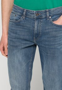 Hombre vestido con jeans de mezclilla azul medio y camisa verde, con la mano izquierda relajada a un lado y la mano derecha en el bolsillo.