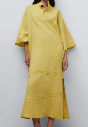 Femme portant une robe jaune ample mi-longue avec des manches larges, un col fendu et des détails de couture sur un fond uni.