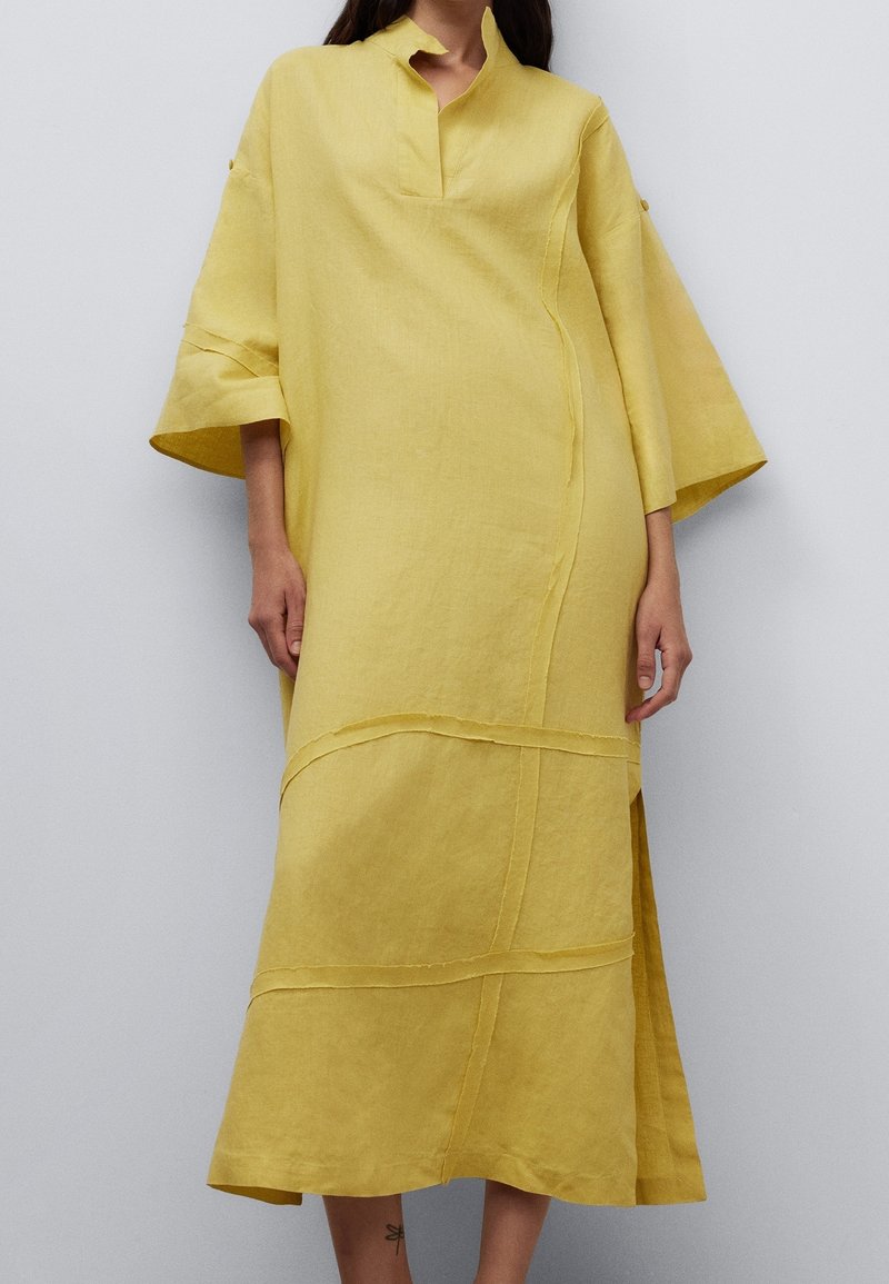 Femme portant une robe jaune ample mi-longue avec des manches larges, un col fendu et des détails de couture sur un fond uni.