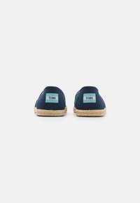 Scarpe slip-on in tela blu navy con suola in corda di juta e due etichette del logo TOMS sul tallone. Texture liscia con forma arrotondata.