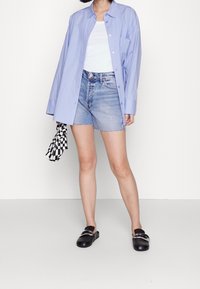 Camisa de manga larga azul claro, camiseta blanca de tirantes, pantalones cortos de mezclilla desgastados, zuecos negros con accesorios plateados, sosteniendo un objeto de cuadros blanco y negro.