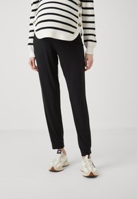 Svarte tapered joggebukser med en glatt tekstur, sammen med en stripete hvit og svart genser med gullknapper.