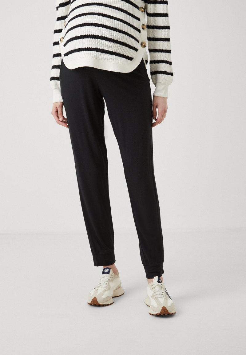 Svarte tapered joggebukser med en glatt tekstur, sammen med en stripete hvit og svart genser med gullknapper.