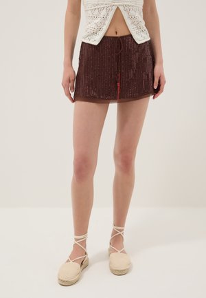 Femme portant un haut en crochet blanc, une mini-jupe marron à sequins avec un cordon et des sandales espadrilles crème avec liens à la cheville, debout sur un sol blanc.