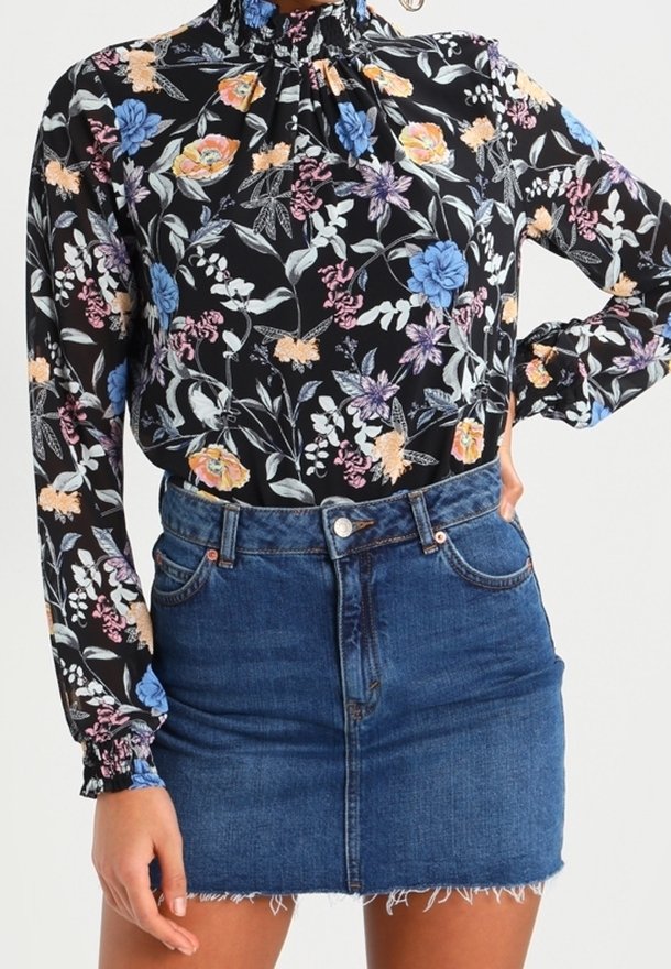 Blouse fleurie noire avec col volanté et manches longues, ornée de fleurs multicolores. Associée à une mini-jupe en denim bleue avec un ourlet effiloché.