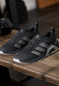 Chaussures de sport noires avec des accents gris, empeigne en mesh texturé, semelle en caoutchouc blanche et design à trois bandes bien visible. Positionnées sur une surface en bois.