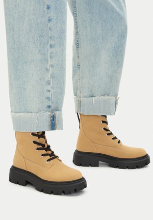 Beżowe botki bojówki wykonane z gładkiej skóry z masywną czarną podeszwą. Posiadają czarne sznurówki i nity, noszone z podwiniętymi jasnoniebieskimi dżinsami.