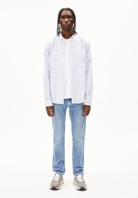 Lange mouwen, lichtblauw en wit gestreept overhemd met knoopjes over een wit T-shirt, gecombineerd met lichtblauwe jeans en grijze sneakers.