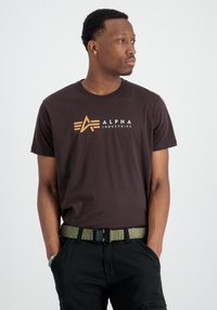 Brūna kokvilnas T-krekls ar baltu un oranžu "Alpha Industries" logotipu. Modelis valkā zaļu jostu un melnus bikses ar kabatām.