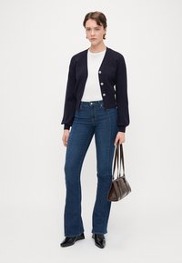 Ναυτικό πουλόβερ με μακριά μανίκια και ασημένια κουμπιά, συνδυασμένο με ψηλόμεσες, καμπάνες σκούρα μπλε τζιν και καφέ τσάντα. Μαύρες μυτερές γόβες.