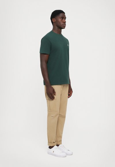T-shirt en coton vert foncé à manches courtes, associé à un pantalon cargo beige avec des poches latérales, et des baskets blanches.