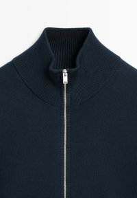 Mörkblå stickad tröja med ribbad hög krage och en silverfärgad dragkedja helt stängd i mitten fram.