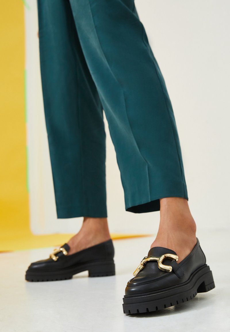Personne portant un pantalon court couleur sarcelle et des mocassins noirs épais avec un grand détail de chaîne dorée sur un sol blanc.