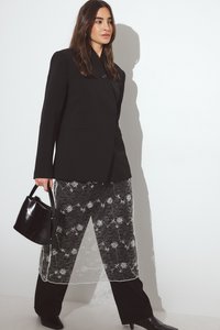 Zwarte op maat gemaakte blazer met enkele knoop, gedragen over een doorschijnende zwarte kanten rok met florale borduur. Accessoires zijn onder andere een kleine zwarte handtas.