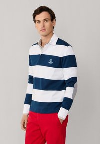 Uomo con una maglia da rugby a righe bianche e blu navy con colletto e pantaloni rossi, in piedi davanti a uno sfondo grigio chiaro uniforme.