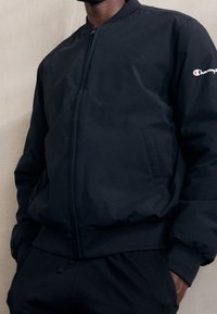 Svart bomberjacka med blixtlås fram och sidofickor. Har ribbade ärmslut och krage, tillverkad av ett slätt, lätt material.