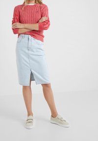 Jupe crayon en denim bleu clair avec une fente à l'avant, associée à un pull rouge en maille torsadée et des chaussures beige à enfiler. Design simple avec un minimum de ferrures.