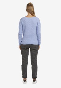 Frau, die mit dem Rücken steht, trägt einen hellblauen, gemusterten Pullover, dunkelgraue Hose, weiße Turnschuhe und hat einen Haarknoten.