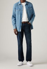 Denim shirt in light blue, white t-shirt, dark jeans, brown belt, and white sneakers. Features chest pockets and button-up design.