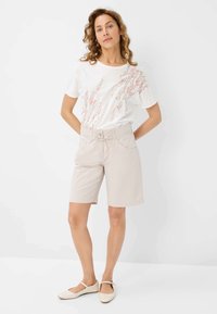 Shorts beiges légers avec une coupe ajustée, associés à un t-shirt blanc arborant un motif floral pastel. Les chaussures sont de couleur claire et plates.