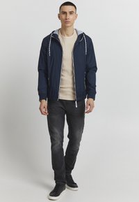 Cazadora de marina con forro de capucha gris, puños y dobladillo elásticos, combinada con un suéter beige y jeans oscuros, llevada con zapatillas negras.