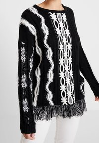 Black and white knitted sweater featuring intricate patterns, textured fabric, and a fringed hem for added detail. Long sleeves included.