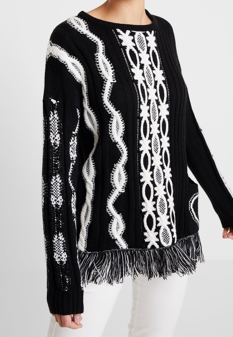 Black and white knitted sweater featuring intricate patterns, textured fabric, and a fringed hem for added detail. Long sleeves included.