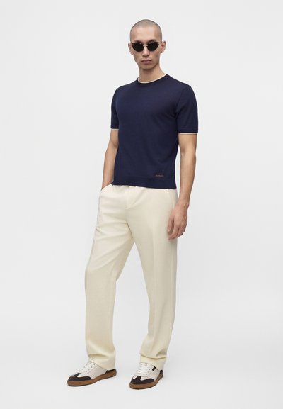 Homme portant une chemise bleu foncé à manches courtes, un pantalon beige, des lunettes de soleil foncées et des baskets bicolores, debout les mains dans les poches.
