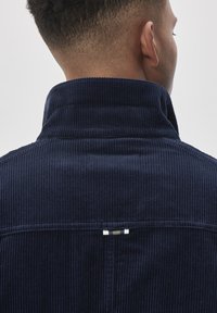 Jachetă din catifea navy cu guler înalt, având o textură ribbed și un detaliu cu etichetă în zona gâtului. Linii curate și o croială bine definită.