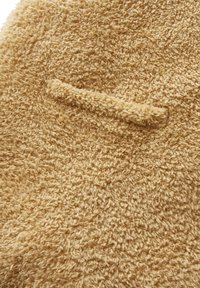 Flauschiger beiger Stoff mit strukturierter Oberfläche; verfügt über eine kleine, auffällige Tasche. Weiche und plüschige Optik mit variierender Fadendicke.