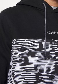 Sudadera negra con un patrón abstracto en blanco y negro texturizado en la mitad inferior. Incluye un logo y cordones de contraste.