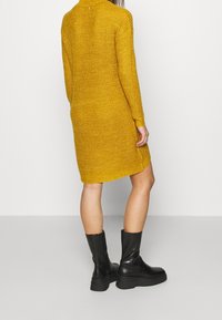 Abito lavorato a maglia giallo senape con maniche lunghe, una vestibilità leggermente oversize e dettagli a costine sul colletto e sull'orlo. Abbinato a stivaletti neri alla caviglia.