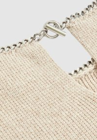 Nahaufnahme eines hellbeigen Strickstoffs mit einem Metalldetail des Reißverschlusses, ausgestattet mit einem runden Ziehgriff, der an einer Kette befestigt ist.