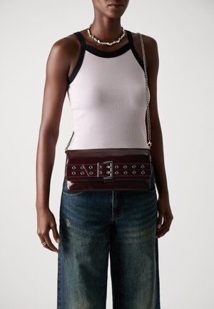 Burgundy leather crossbody bag with silver hardware, featuring grommets, a buckle accent, and a chain strap. Worn over a light tank top.