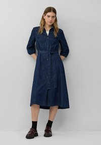 Denim jurkjuhl, donkerblauw, knoopsluiting aan de voorkant, driekwart mouwen, ceintuur in de taille, knielengte, recht gesneden, gedragen met donkere schoenen en sokken.