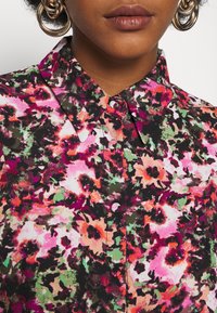 Blouse florale au motif multicolore avec des tons roses, verts et noirs. Design avec col, fermeture à boutons et texture de tissu douce.