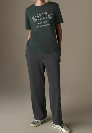 Person iført mørkegrøn SOHO Yoga Studio t-shirt, løstsiddende sorte bukser og sølvfarvede sneakers, stående med hænderne i lommerne.