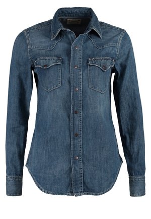 Persona que lleva una bufanda marrón, camisa vaquera azul con las mangas remangadas, pantalones negros y botines marrones de tacón alto, de pie sobre un fondo liso.