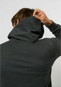 Hanorac gri cu o textură moale, având un glugă cu șnur și o croială confortabilă. Pe spate se vede o mână ajustând gulerul.