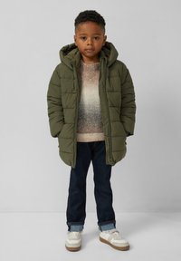 Kind, der eine grüne Steppjacke, einen mehrfarbigen Pullover, dunkle Jeans mit Aufschlägen und weiße Sneaker trägt, steht vor einem schlichten Hintergrund.
