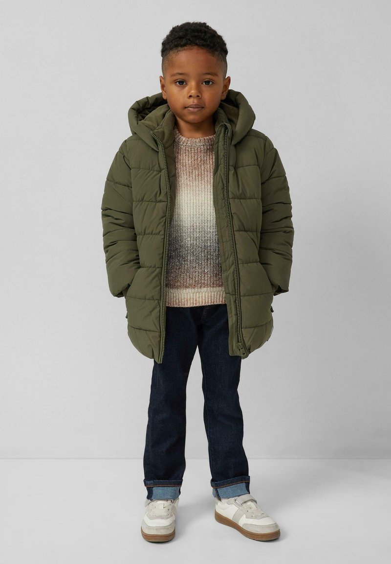 Kind, der eine grüne Steppjacke, einen mehrfarbigen Pullover, dunkle Jeans mit Aufschlägen und weiße Sneaker trägt, steht vor einem schlichten Hintergrund.