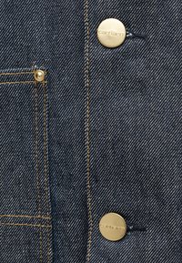 Σκούρο μπλε ύφασμα τζιν με διαγώνιο ύφανση, με χρυσά μεταλλικά κουμπιά και αντίθετη κίτρινη ραφή κατά μήκος των τσεπών και των ραφών.