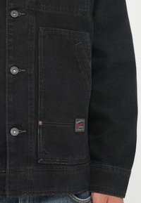 Black denim jacket with front buttons, chest pocket, and brand patch. Smooth texture, tailored cut, and stitched detailing on the hem.