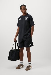 T-shirt nero adidas con logo, pantaloni corti neri, calze con righe, scarpe marroni e una borsa a tracolla nera, indossato da una figura maschile.