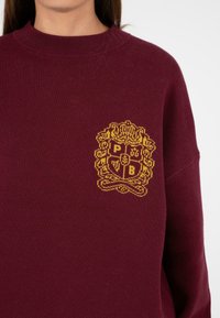 Burgundowy sweter dzianinowy z teksturowanym żółtym emblematem z inicjałami "P", "B" oraz wzorem tarczy na klatce piersiowej. Okrągły dekolt.