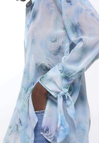 Sheer, light blue blouse with a floral pattern, ties at the cuffs, slight shimmer, loose fit, featuring an angled side slit.