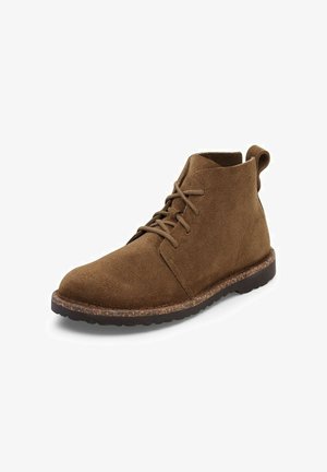 Stivale in suede marrone alla caviglia con punta rotonda, chiusura con lacci, superficie testurizzata e suola in sughero contrastante per una maggiore aderenza e durata.