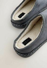 Szare zamszowe kapcie z miękkim białym futerkiem w środku, plecionym czarno-białym wykończeniem oraz metką wewnątrz z napisem "NEXT". Płaski, wsuwany fason.