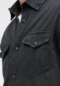 Camicia nera con bottoni, realizzata in cotone, con colletto classico, due tasche sul petto con patta e un'etichetta del logo gialla sulla tasca sinistra.